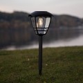 Czarna solarna lampa ogrodowa wbijana w ziemię, z przezroczystym kloszem i ciepłym światłem LED, ustawiona na trawie nad jeziorem o zmierzchu.