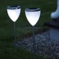 Dwie białe lampy solarne wbijane w trawę przy żwirowej nawierzchni, świecące chłodnym białym światłem po zmroku.