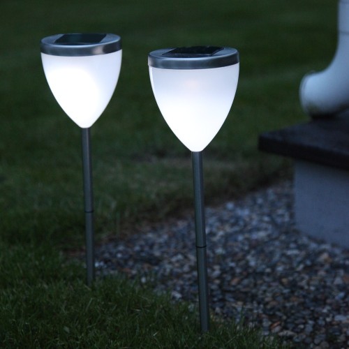 Dwie białe lampy solarne wbijane w trawę przy żwirowej nawierzchni, świecące chłodnym białym światłem po zmroku.