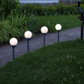 Cztery solarne lampy ogrodowe z mlecznymi kulami ustawione wzdłuż ścieżki na trawniku, dekoracyjne oświetlenie zewnętrzne LED wbijane do gruntu.
