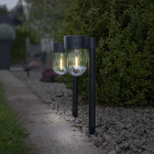 Dwie czarne solarne lampy ogrodowe na słupkach z przezroczystymi kloszami i widocznym źródłem światła, ustawione wzdłuż betonowej ścieżki przy pasie żwiru, na tle zielonego żywopłotu.