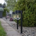Dwie czarne solarne lampy ogrodowe z przezroczystymi kloszami i widocznym światłem, ustawione wzdłuż betonowej ścieżki przy pasie żwiru, na tle zielonego żywopłotu.