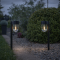 Dwie czarne solarne lampy ogrodowe z przezroczystymi kloszami i widocznym światłem, ustawione po obu stronach betonowej ścieżki przy pasie żwiru, na tle trawnika i zielonego żywopłotu o zmierzchu.