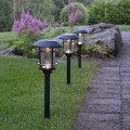Trzy czarne solarne lampy ogrodowe z przezroczystymi kloszami osłoniętymi kratką, ustawione w trawie przy ścieżce z kostki brukowej, na tle roślin i dużego kamienia.