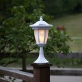 Solarna biała lampa ogrodowa, ustawiona na drewnianym słupku przy ogrodzeniu, na tle zielonych drzew, z widocznym światłem wewnątrz klosza.