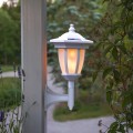 Solarna biała lampa ogrodowa zamocowana do drewnianego słupka przy ogrodzeniu, na tle roślin i ścieżki, z widocznym światłem wewnątrz klosza.