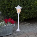 Solarna biała lampa ogrodowa ustawiona na kamiennej nawierzchni obok metalowej donicy z czerwonymi kwiatami, na tle zielonych krzewów, z widocznym światłem wewnątrz klosza.