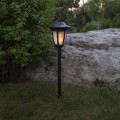 Solarna czarna lampa ogrodowa wbijana w ziemię, emitująca ciepłe światło, ustawiona na trawie obok dużego kamienia i zielonych krzewów.