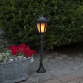 Czarna solarna lampa ogrodowa w formie latarenki ustawiona na kamiennej nawierzchni obok metalowej donicy z czerwonymi kwiatami, świecąca ciepłym światłem na tle gęstych zielonych krzewów o zmierzchu.