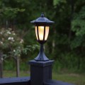 Czarna solarna lampa ogrodowa w formie latarenki zamontowana na słupku drewnianego ogrodzenia, świecąca ciepłym światłem na tle zielonych drzew o zmierzchu.