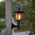 Czarna solarna lampa ogrodowa zamontowana na ścianie budynku, świecąca ciepłym światłem na tle roślin i ogrodu o zmierzchu.