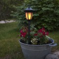 Solarna czarna lampa ogrodowa ustawiona w metalowej donicy z czerwonymi i białymi kwiatami, stojąca na trawniku na tle zielonych krzewów o zmierzchu.