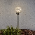 Lampa solarna ogrodowa kula LED w kolorze srebrnym z dekoracyjnym, pękanym kloszem, emitująca ciepłe białe światło. Zamontowana w ziemi przy ścianie budynku, w otoczeniu roślin i kory ogrodowej.