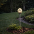 Solarna lampa ogrodowa z kulistym kloszem z efektem spękanego szkła, emitująca ciepłe białe światło, zamontowana na metalowym słupku na trawniku przy rabacie.