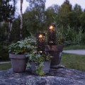 Dwie czarne solarne lampy ogrodowe LED z ażurową obudową ustawione w donicach na kamiennej powierzchni, emitujące ciepłe białe światło i tworzące dekoracyjny nastrój w ogrodzie o zmierzchu.