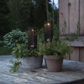 Dwie czarne solarne lampy ogrodowe z ażurowym wzorem, ustawione w donicach z zielonymi roślinami na drewnianym stole na tarasie, emitujące ciepłe światło po zmroku.