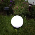 Lampa solarna ogrodowa z białą kulą ustawiona w trawie wśród roślin i kwiatów, z oddzielnym panelem solarnym widocznym w tle, tworząca subtelne oświetlenie dekoracyjne w ogrodzie.