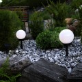 Lampy solarne ogrodowe z mlecznymi kulami na czarnych słupkach umieszczone wśród jasnych kamieni i zielonych roślin, emitujące delikatne światło dekoracyjne w przydomowej aranżacji ogrodu.