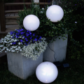 Białe kule solarne LED umieszczone wśród kwiatów w betonowych donicach oraz na trawie, emitujące delikatne światło i tworzące dekoracyjny efekt świetlny w ogrodowej aranżacji po zmroku.