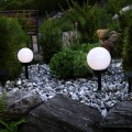 Dwie lampy solarne ogrodowe w kształcie białych kul na czarnych słupkach ustawione wśród kamieni i zielonych roślin, delikatnie oświetlające rabatę.