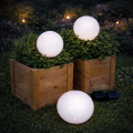 Białe kule solarne LED ustawione w drewnianych skrzyniach z roślinami oraz na trawie, emitujące delikatne światło i tworzące nastrojową aranżację ogrodową po zmroku.