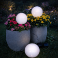 Białe kule solarne LED umieszczone w dużych donicach z kolorowymi kwiatami oraz na trawie, emitujące delikatne światło i tworzące nastrojową dekorację ogrodową po zmroku.