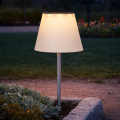 Solarna lampa ogrodowa LED z białym kloszem w kształcie abażuru i stalowym słupkiem, zamontowana na rabacie przy trawniku i kamiennej obręczy, dekoracyjne oświetlenie zewnętrzne do ogrodu i przy ścieżkach.