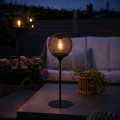 Solarna lampa ogrodowa LED z czarną ażurową osłoną i widoczną żarówką ustawiona na stoliku tarasowym przy sofie i donicach z kwiatami, dekoracyjne oświetlenie zewnętrzne do ogrodu i na patio.