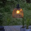 Solarna lampa ogrodowa LED z czarną ażurową osłoną i widoczną żarówką zawieszona na przewodzie na tle zieleni i roślin w donicach.