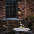 Solarna lampa ogrodowa LED z czarną ażurową osłoną i widoczną żarówką zawieszona na przewodzie przy ceglanej elewacji obok okna, dekoracyjne oświetlenie zewnętrzne przy stoliku i krzesłach.