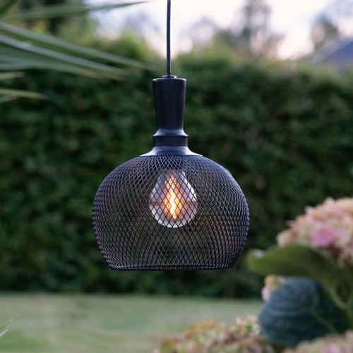 Solarna lampa ogrodowa LED z czarną ażurową osłoną i widoczną żarówką zawieszona na przewodzie na tle trawnika i roślin, dekoracyjne oświetlenie zewnętrzne do ogrodu.