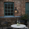 Solarna lampa ogrodowa LED z czarną ażurową osłoną i widoczną żarówką zawieszona na przewodzie przy ceglanej ścianie obok okna, dekoracyjne oświetlenie zewnętrzne nad stolikiem i krzesłami.