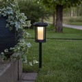 Solarna lampa ogrodowa LED na czarnej sztycy wbitej w trawnik obok donicy z zieloną rośliną. Oprawa z przydymionym kloszem emituje ciepłe światło, oświetlając przestrzeń w ogrodzie przy ścieżce.