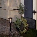 Solarna lampa ogrodowa LED na czarnej sztycy wbitej w żwir obok dużej donicy z zieloną rośliną. Oprawa z przydymionym kloszem emituje ciepłe światło, oświetlając przestrzeń przy jasnej drewnianej powierzchni.
