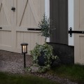 Solarna lampa ogrodowa LED na czarnej sztycy wbitej w żwir obok dużej donicy z zieloną rośliną. Oprawa z przydymionym kloszem emituje ciepłe światło, ustawiona przy jasnych drewnianych drzwiach z widocznymi zawiasami.