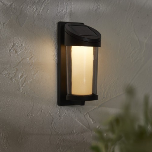Solarny kinkiet ścienny LED z czarną obudową i lekko przydymionym szklanym kloszem, emitujący ciepłe białe światło, zamontowany na jasnej elewacji budynku jako oświetlenie zewnętrzne przy wejściu lub na tarasie.