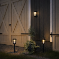 Solarne lampy ogrodowe LED z czarną obudową i lekko przydymionym kloszem, w tym kinkiet ścienny oraz dwie lampy na słupkach, emitujące ciepłe białe światło przy budynku.
