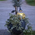 Solarny reflektor ogrodowy LED zamontowany w donicy z roślinami, z głowicą świetlną i panelem solarnym osadzonymi na giętkich ramionach, emitujący ciepłe białe światło podkreślające zieleń.