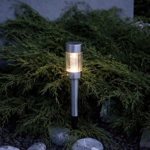 Solarna lampa ogrodowa LED na szpikulcu z przezroczystym kloszem i metalową obudową, emitująca ciepłe światło jako dekoracyjne oświetlenie rabat i krzewów w ogrodzie.