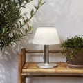 Solarna ogrodowa lampka stołowa LED z białym kloszem i metalową podstawą, ustawiona na drewnianej półce w otoczeniu roślin – nastrojowe oświetlenie ogrodu, tarasu lub balkonu.