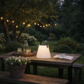 Solarna ogrodowa lampa LED z białym kloszem i uchwytem, ustawiona na drewnianym stole w ogrodzie po zmroku, tworząca nastrojowe oświetlenie wśród roślin i girlandy świetlnej.