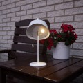 Biała lampa stołowa z okrągłą podstawą i kloszem z uchwytem, emitująca ciepłe światło, ustawiona na drewnianym stole przy ceglanej ścianie, obok donicy z czerwonymi kwiatami, wyposażona w zwisający sznurek z włącznikiem.