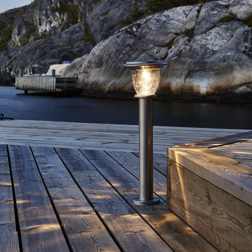 Solarna srebrna lampa ogrodowa słupek z przezroczystym kloszem emitującym ciepłe światło, zamontowana na drewnianym pomoście nad wodą, na tle skalistego brzegu i niewielkiego pomostu.