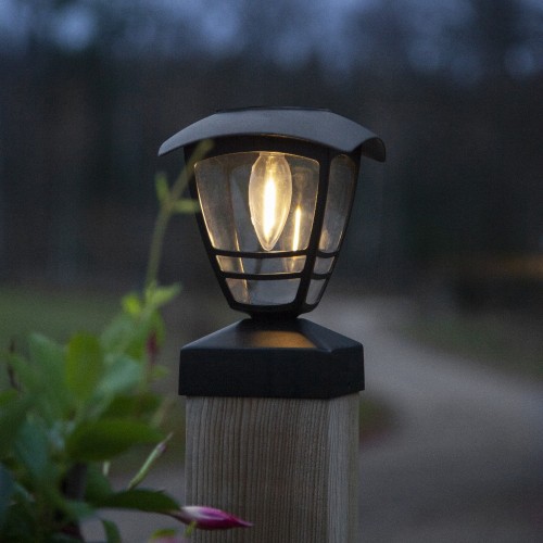 Czarna solarna lampa ogrodowa z dekoracyjnym kloszem emitującym ciepłe światło, zamontowana na drewnianym słupku przy ścieżce ogrodowej, na tle rozmytej zieleni i roślin.