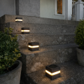 Cztery szare solarne lampy ogrodowe o kostkowym kształcie emitujące ciepłe światło, ustawione na kamiennych schodach przy wejściu do domu, w otoczeniu donic z zielonymi roślinami.