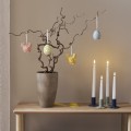 Porcelanowe dekoracje wielkanocne LED w kształcie kurczaczków i jajek zawieszone na gałązkach w wazonie, pastelowe ozdoby do wnętrz z ciepłym światłem ustawione na drewnianym stole obok świec.