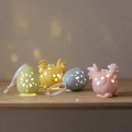 Porcelanowe dekoracje wielkanocne LED w kształcie kurczaczków i jajek w pastelowych kolorach ustawione na drewnianej półce, ozdoby z ażurowymi wycięciami emitujące ciepłe światło.