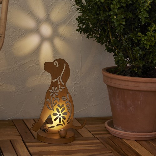 Solarna lampa ogrodowa LED w kształcie psa z dekoracyjnym wycinanym wzorem, ustawiona na drewnianym tarasie przy donicy z rośliną. Ozdoba solarna z podświetleniem tworząca efekt wzoru na ścianie, dekoracja do ogrodu, na taras lub balkon.