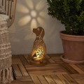 Solarna lampa ogrodowa LED w kształcie psa z dekoracyjnym wycinanym wzorem, ustawiona na drewnianym tarasie przy donicy z rośliną. Ozdoba z podświetleniem tworząca efekt wzoru na ścianie, dekoracja do ogrodu, na taras lub balkon.