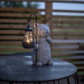 Solarna figurka ogrodowa LED w kształcie kota trzymającego latarenkę na haczyku, ustawiona na metalowym stoliku. Dekoracja solarna do ogrodu, na taras lub balkon, z podświetlaną latarnią tworzącą efekt świetlny.